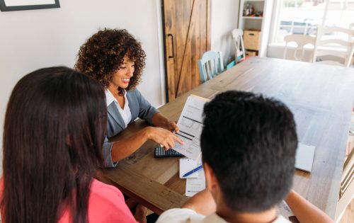 A female financial advisor sits with a millennial age married couple at a dining room table with laptop and financial reports helping with their monthly budget and investments. She is showing her clients how to read financial reports and monthly finances, pay taxes and save money for the future.